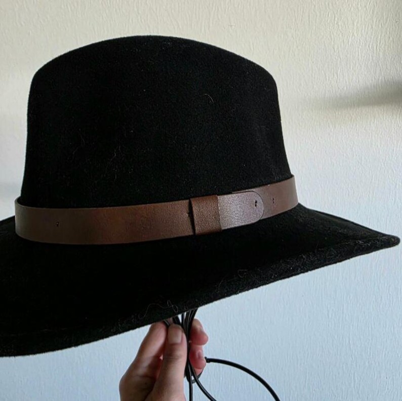 Pu&ograve; includere: Un cappello fedora nero caratterizzato da un'ampia fascia marrone liscia che circonda la corona. Il cappello presenta una parte superiore strutturata e arrotondata e una tesa larga e piatta. Sottili cordini neri pendono dalla tesa.