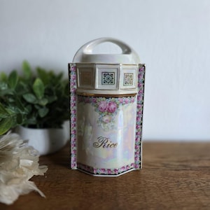 Pode incluir: Um pote de arroz de porcelana vintage com uma alça, com um acabamento perolado. O pote é decorado com rosas cor-de-rosa e a palavra "Rice" em escrita elegante. As bordas são aparadas com um padrão floral e detalhes dourados.