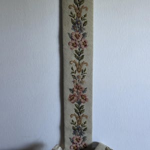 May include: A long, narrow, embroidered wall hanging with a floral design in pink, blue, and green on a beige background. The top has a decorative gold-colored trim. A matching small piece and a gold box are also visible.