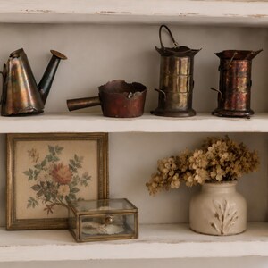 May include: A white wooden shelf containing a collection of items. The top shelf holds a metallic watering can, a small copper pan, and two cylindrical metal containers. The bottom shelf features a framed floral print, a glass box, and a ceramic vase.