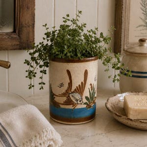 May include: A cylindrical ceramic planter with a cream-colored base, featuring a hand-painted brown fish motif, green reeds, and a horizontal blue band near the bottom. The planter contains a small, leafy green plant with thin, trailing stems.