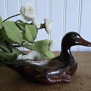 Op de afbeelding: Een bruin en zwart keramieken eendenbeeldje met een rode snavel. De eend staat op een houten oppervlak, met witte bloemen en groene bladeren op de achtergrond. De eend is een decoratief item.