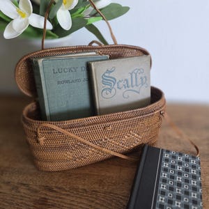 Może przedstawiać: Brązowa, pleciona torebka z rattanu z uchwytem, ​​zawierająca dwie stare książki. Jedna książka nosi tytuł "Lucky Dog", a druga "Scally". Trzecia książka z czarną okładką i geometrycznym wzorem leży obok torby.