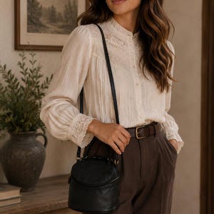 May include: A cream-coloured long-sleeve blouse with a high collar, button-down front, and lace detailing on the chest and cuffs. The blouse is paired with dark brown trousers featuring a brown leather belt and a small black round crossbody bag.