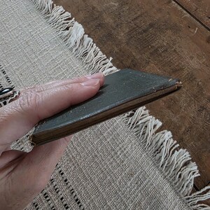 Puede incluir: Un peque&ntilde;o libro antiguo con una cubierta verde oscuro. El libro se sostiene en una mano, mostrando sus bordes desgastados y su apariencia envejecida. El libro descansa sobre un mantel individual beige con flecos.
