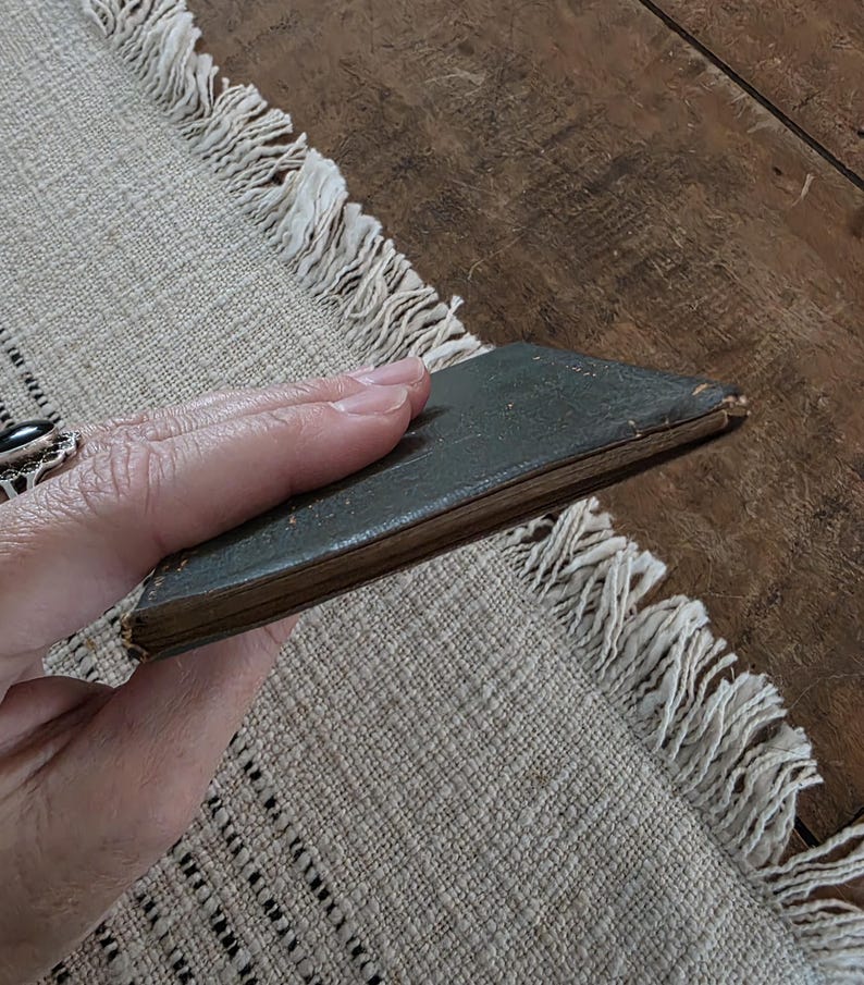 Puede incluir: Un peque&ntilde;o libro antiguo con una cubierta verde oscuro. El libro se sostiene en una mano, mostrando sus bordes desgastados y su apariencia envejecida. El libro descansa sobre un mantel individual beige con flecos.