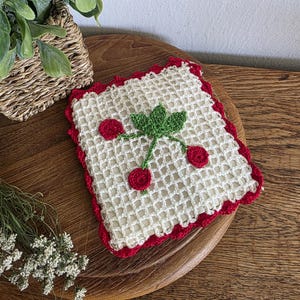 Puede incluir: Un posavasos cuadrado de ganchillo blanco roto con un borde festoneado rojo. Presenta una hoja verde y tres aplicaciones de cerezas rojas. El posavasos está colocado sobre una superficie de madera, con una cesta y vegetación en el fondo.