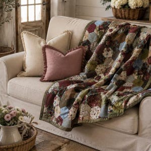 Puede incluir: Una colcha de patchwork con un patrón hexagonal en varios tonos de burdeos, verde oliva, crema y azul claro. La colcha está colocada sobre un sofá de color crema, que también cuenta con un cojín de color crema liso y un cojín rosa palo liso.