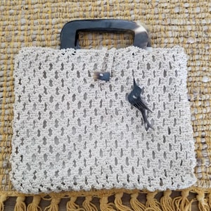 May include: Cream-colored macrame tote bag with a dark handle and a decorative bird-shaped charm. The bag features a woven pattern and a button closure. The bag is sitting on a textured, yellow and beige surface.
