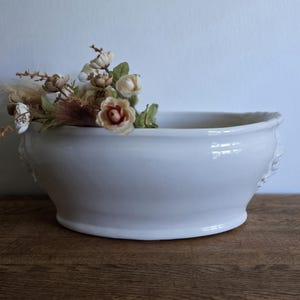 Puede incluir: Un cuenco de cerámica blanco con forma ovalada. El cuenco tiene asas decorativas y está lleno de flores secas y vegetación. El cuenco está sobre una superficie de madera. El cuenco mide unos 30 cm de largo.