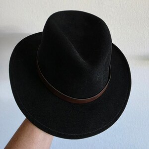 Pu&ograve; includere: Un cappello fedora nero caratterizzato da una corona strutturata con una piega centrale e una tesa larga e piatta. Il cappello &egrave; arricchito da una sottile fascia in pelle marrone liscia che circonda la base della corona, creando un netto contrasto.
