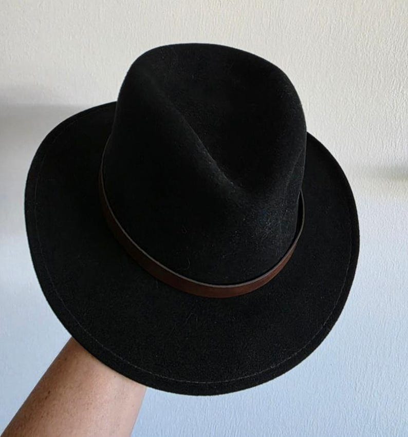 Pu&ograve; includere: Un cappello fedora nero caratterizzato da una corona strutturata con una piega centrale e una tesa larga e piatta. Il cappello &egrave; arricchito da una sottile fascia in pelle marrone liscia che circonda la base della corona, creando un netto contrasto.