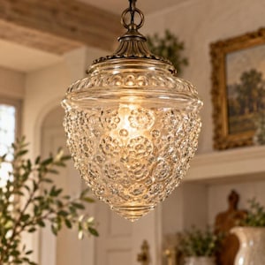 Puede incluir: Una lámpara colgante con una pantalla de vidrio transparente texturizado con un patrón floral repetitivo. La pantalla tiene forma de bellota y está suspendida de una tapa decorativa de metal en tono latón antiguo con una anilla de montaje a juego.