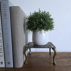 May include: A small, decorative brass plant stand with a white ceramic pot holding a green leafy plant. The stand has ornate detailing and curved legs. Books are visible in the background.