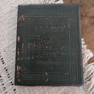 Puede incluir: Un libro vintage de color verde oscuro con el t&iacute;tulo "ESSAYS" y el nombre del autor "EMERSON" en relieve en la portada. El libro tiene un aspecto desgastado y texturizado, con detalles dorados y bordes decorativos. El libro est&aacute; sobre una superficie texturizada blanca y beige.