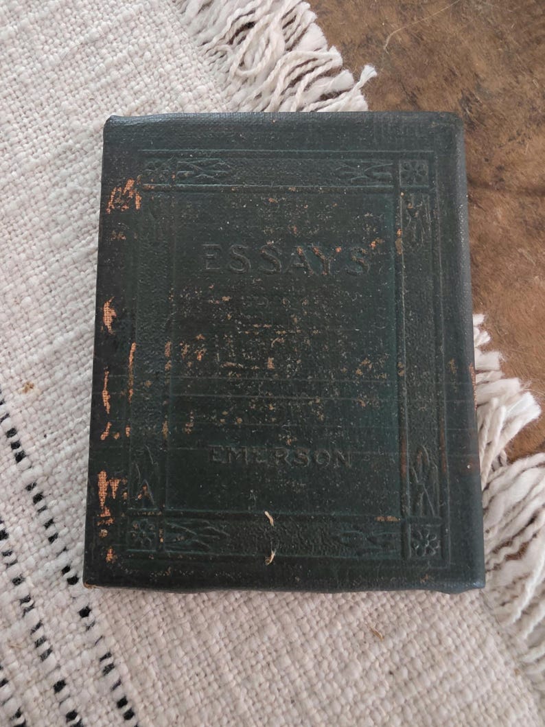 Puede incluir: Un libro vintage de color verde oscuro con el t&iacute;tulo "ESSAYS" y el nombre del autor "EMERSON" en relieve en la portada. El libro tiene un aspecto desgastado y texturizado, con detalles dorados y bordes decorativos. El libro est&aacute; sobre una superficie texturizada blanca y beige.