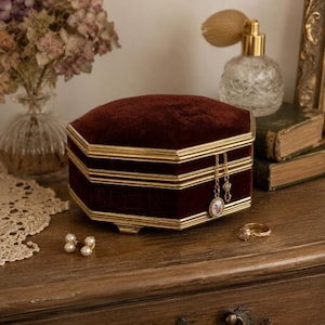 May include: A hexagonal jewelry box covered in deep burgundy velvet with gold-toned metal trim. The box features a hinged lid and a small gold chain with a floral pendant hanging from the front edge. It sits alongside pearl earrings and a gold ring.