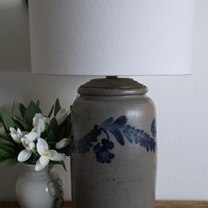 Puede incluir: Lámpara de gres con pantalla de tela blanca. La base de la lámpara es de color gris azulado con un diseño floral azul. La lámpara se asienta sobre una base negra. Un jarrón de flores blancas está al lado de la lámpara.