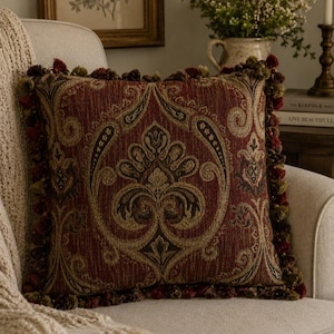 May include: A square throw pillow featuring a deep red and gold damask pattern with intricate scrollwork. The edges are finished with a dense fringe of small, multi-colored tassels in shades of burgundy, olive green, and gold, creating a textured border.