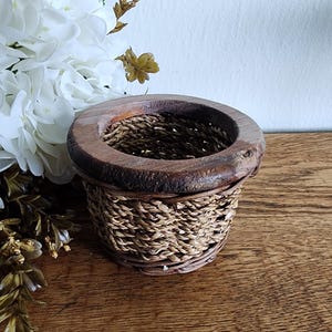 Może przedstawiać: Mały, ręcznie wykonany kosz z tkanym, cylindrycznym korpusem i grubą, okrągłą drewnianą obręczą. Kosz wykonany jest z naturalnych włókien i drewna, o rustykalnej estetyce. Kosz stoi na drewnianej powierzchni.