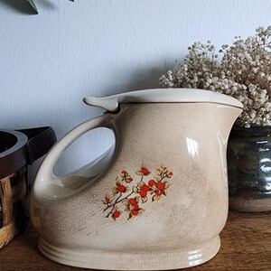 Puede incluir: Una jarra de cerámica beige con tapa y asa grande. La jarra presenta un diseño floral con bayas rojas y detalles amarillos. La jarra está sobre una superficie de madera.