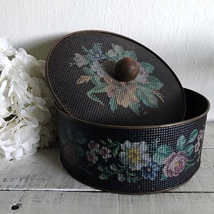 May include: A black metal storage container with a floral design. The lid is slightly ajar, revealing the interior. The container features a cross-stitch floral pattern in pastel colors. A wooden knob sits atop the lid. The container is on a white wooden surface.