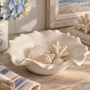 Pode incluir: Uma tigela de cerâmica na cor creme com uma borda ondulada e festonada. A tigela contém uma coleção de conchas naturais e um pedaço de coral branco. A superfície da tigela é lisa e brilhante, refletindo a luz em suas bordas curvas.