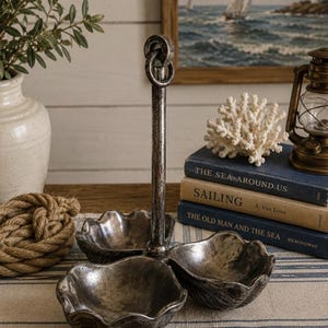May include: A three-section metal serving tray with a dark, weathered silver finish and a central vertical handle topped with a ring. The tray features three shell-shaped bowls arranged around the central post, creating a tiered display piece.