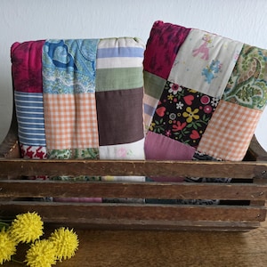May include: Two patchwork pillows in a rustic wooden crate. The pillows feature a variety of fabric patterns, including stripes, gingham, floral, and paisley. The crate is made of weathered wood, adding to the vintage aesthetic. Yellow flowers are in the foreground.