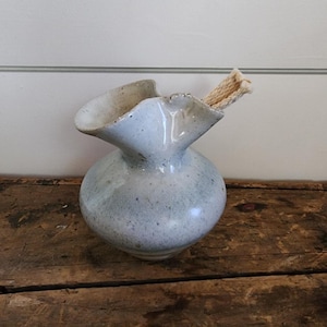 May include: A speckled, light blue ceramic vase with a unique, ruffled opening. The vase has a rounded body and contains a bundle of beige, rope-like material. The vase sits on a rustic, wooden surface.