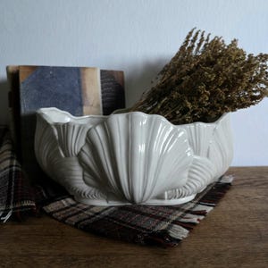 Könnte beinhalten: Weißer Keramik-Pflanztopf in Muschelform mit glänzendem Finish. Der Pflanztopf ist mit getrockneten braunen Pflanzen gefüllt und steht auf einem karierten Stoff. Im Hintergrund ist ein Stapel Bücher zu sehen.