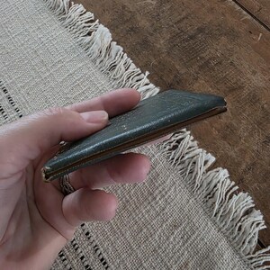 Puede incluir: Una peque&ntilde;a cartera rectangular de cuero verde oscuro con aspecto desgastado. La cartera se sostiene en una mano y parece estar cerrada. Los bordes son de color marr&oacute;n claro. El fondo es un mantel individual beige con flecos.