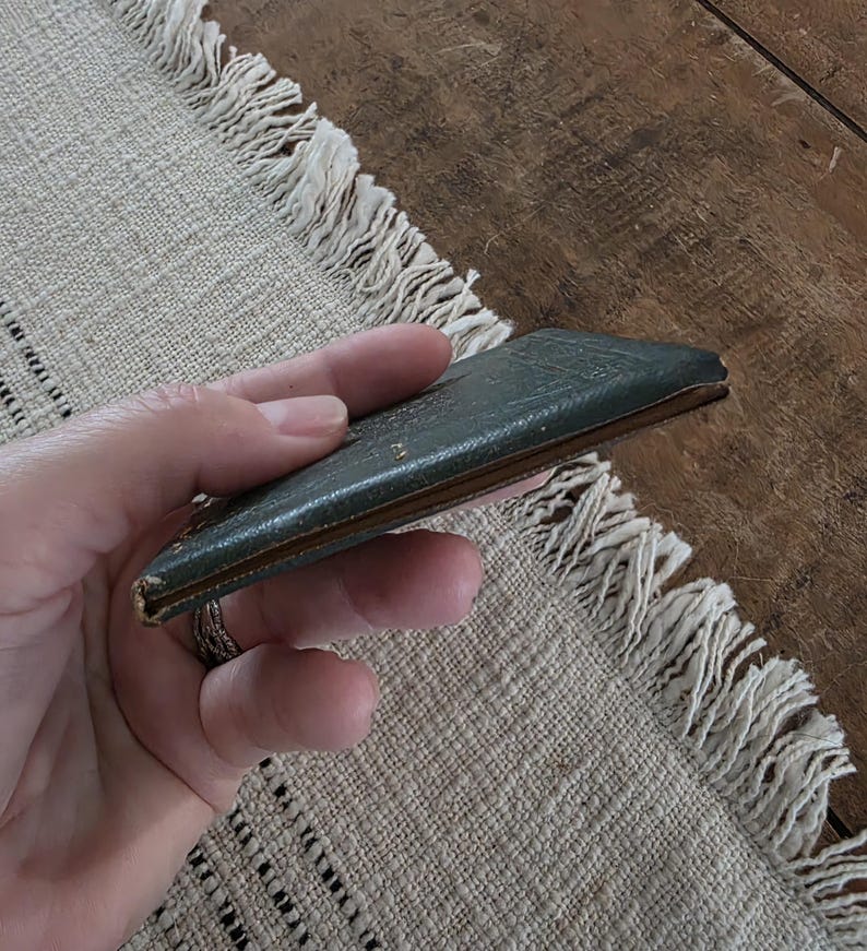 Puede incluir: Una peque&ntilde;a cartera rectangular de cuero verde oscuro con aspecto desgastado. La cartera se sostiene en una mano y parece estar cerrada. Los bordes son de color marr&oacute;n claro. El fondo es un mantel individual beige con flecos.