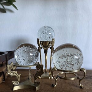 May include: Three clear glass spheres with air bubbles are displayed on ornate brass stands. The stands have decorative details and varying designs. The spheres are on a wooden surface.
