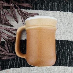 Pode incluir: Uma caneca de cerâmica artesanal com exterior castanho e borda branca. A caneca tem uma alça grande e uma textura salpicada. A caneca está sobre uma superfície listrada em preto e branco.