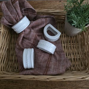 Puede incluir: Una bandeja de mimbre tejida contiene una servilleta de lino marrón doblada y cuatro servilleteros de cerámica blanca. Una pequeña planta en maceta con follaje verde se encuentra en la esquina. Sobre una superficie de madera.