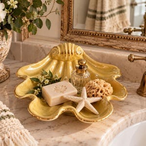 Puede incluir: Un plato de cerámica amarillo brillante con forma de concha que contiene una pastilla de jabón rectangular con una impresión de hoja, un frasco de vidrio transparente con tapón dorado, una esponja natural y una rama de flores blancas.