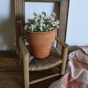 May include: A small terracotta pot filled with white flowers sits on a miniature wooden chair with a woven seat. A rustic wooden picture frame is in the background. A red and white checkered cloth is draped on the side.
