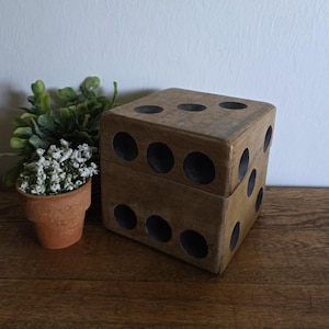 May include: A large, wooden die with black dot markings. The die is brown and sits on a wooden surface. A small terracotta pot with a green plant and white flowers is to the left of the die.