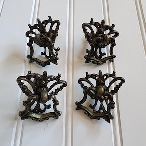 Puede incluir: Cuatro tiradores de cajón antiguos de bronce con diseños ornamentados de volutas. Cada tirador presenta un pomo central y adornos decorativos, ideales para añadir un toque vintage a los muebles. Los tiradores están dispuestos sobre un fondo blanco con listones verticales.