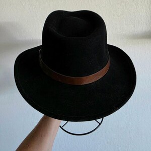 Pu&ograve; includere: Un cappello fedora nero caratterizzato da una corona strutturata e una tesa larga. Il cappello &egrave; arricchito da una fascia in pelle liscia marrone scuro che circonda la base della corona, creando un netto contrasto con il materiale nero.