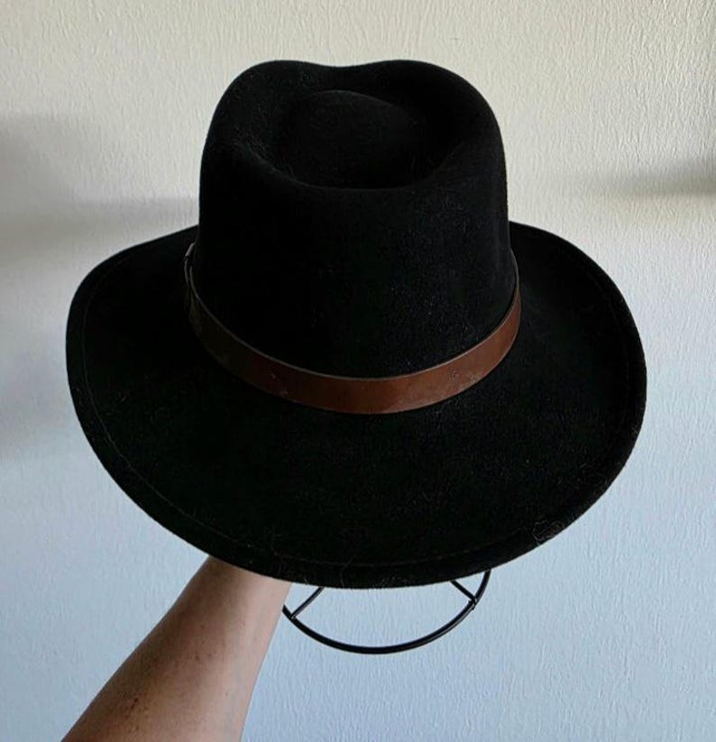 Pu&ograve; includere: Un cappello fedora nero caratterizzato da una corona strutturata e una tesa larga. Il cappello &egrave; arricchito da una fascia in pelle liscia marrone scuro che circonda la base della corona, creando un netto contrasto con il materiale nero.