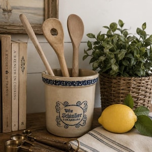 Puede incluir: Una vasija de cerámica beige con un borde decorativo azul y texto azul que dice Vite Schintler STRASBURG. La vasija contiene tres cucharas de madera. Cerca hay tazas medidoras de latón, un limón amarillo y un paño de lino a rayas.