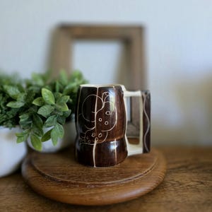 May include: A brown ceramic mug with a white handle and a carved design. The mug features a dark brown exterior with a lighter brown interior and handle. The carved design appears to be an abstract floral or organic pattern.