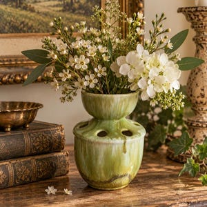 May include: A green ceramic flower frog vase with a multi-hole top, featuring a glossy drip glaze finish in shades of sage and olive. The vase is filled with a bouquet of small white flowers and green leaves, creating a floral arrangement.