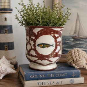May include: A ceramic planter with a glossy white and dark red crackle glaze finish, featuring a central circular medallion depicting a small fish. The vessel has a flared rim and a pedestal base, containing a green leafy plant.