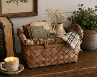 May include: A rectangular woven brown wood basket with two handles, containing a small green book titled The Country Diary, a vintage photograph, and a small vase with dried flowers. A plaid cloth is draped over the edge of the basket.