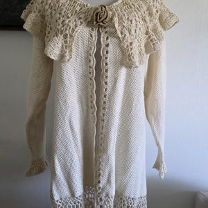 May include: Cream-colored, long-sleeved cardigan with a decorative crochet collar and hem. The open-front sweater features a floral brooch and intricate crochet detailing. The garment is displayed on a mannequin, showcasing its delicate texture and design.