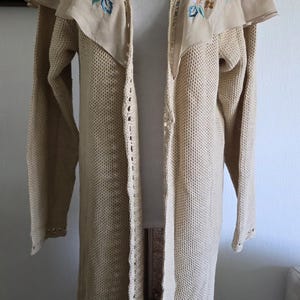 May include: A long, open-front cardigan in a light beige color. The cardigan features a decorative collar with embroidered floral details in pink, green, and blue. The bottom hem has a crocheted ruffle. The garment is displayed on a mannequin.