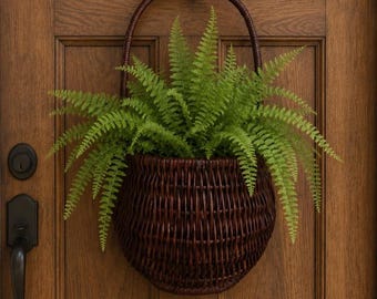 May include: A dark brown woven wicker basket with a tall arched handle hanging from a black metal hook. The basket is filled with a lush, vibrant green fern plant featuring delicate, feathery fronds that spill over the rounded edges.